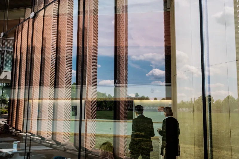 Two people reflected in a glass façade mirroring sky and trees